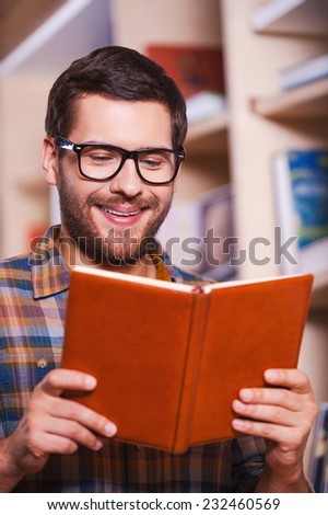 Reading his favorite book. Handsome young man reading book and smiling while standing in front of the bookshelf
