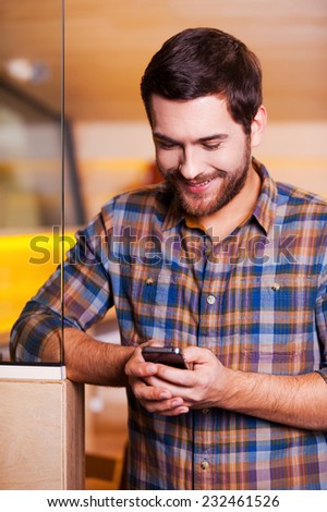 Handsome man texting. Handsome young man looking at his mobile phone and smiling while standing indoors