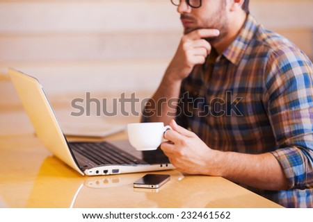 Just inspired. Cropped image of thoughtful young man working on laptop and drinking coffee while sitting in coffee shop