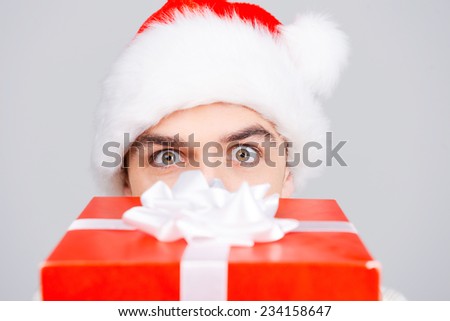 No way! Surprised young man in Santa hat hiding part of his face behind gift box while standing against grey background
