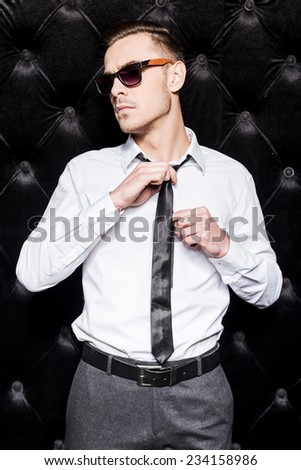 Now I am ready to party. Handsome young man in shirt and tie adjusting his necktie and looking away while standing against black background