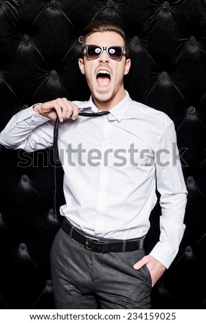 Work hard play hard! Handsome young man in sunglasses taking off his necktie and shouting while standing against black background