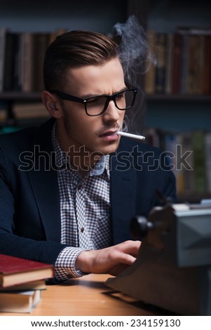 Author at work. Confident young author working at the typewriter and smoking cigarette while sitting at his working place with bookshelf in the background