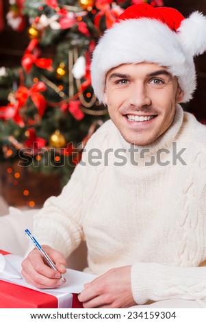 Present for her. Handsome young man in Santa hat writing Christmas letter and smiling with Christmas Tree in the background