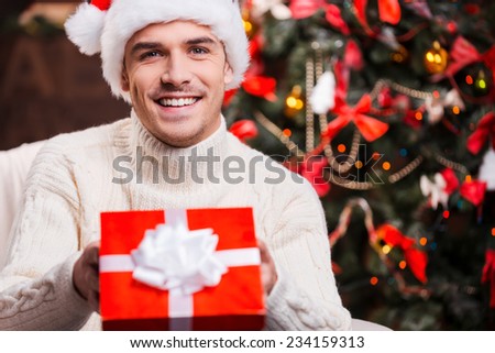 This is for you! Handsome young man in Santa hat stretching out a gift box and smiling with Christmas Tree in the background