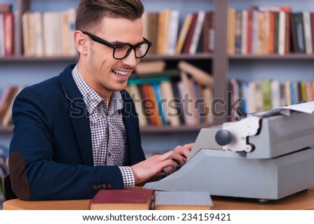 Successful author. Side view of happy young author typing something at the typewriter and smiling while sitting at his working place with bookshelf in the background