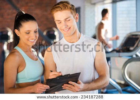 Planning her time in gym. Handsome young male instructor standing close to beautiful woman and showing something at his clipboard while people running on treadmill in the background
