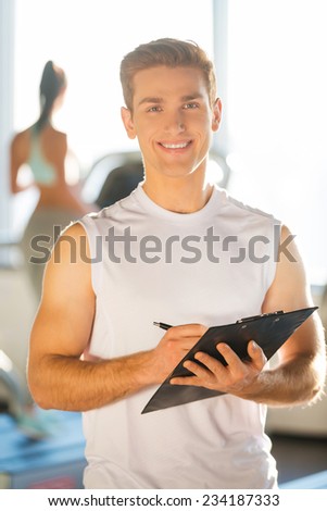 Confident instructor. Handsome young man holding clipboard and smiling while woman running on treadmill in the background