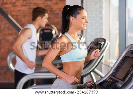 Cardio training. Side view of beautiful young woman running on a treadmill and smiling with man in the background