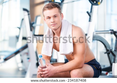 Having a break after workout. Handsome young muscular man holding bottle with water and looking at camera while having a rest in gym
