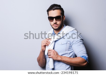Casual cool. Handsome young man in blue shirt tying sweater on her shoulders while standing against grey background