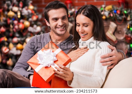 What a great surprise! Beautiful young woman opening a gift box and smiling while her boyfriend sitting close to her on the couch with Christmas decoration in the background