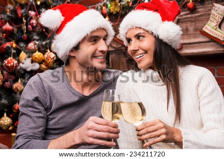 Cheers! Happy young loving couple in Santa hats bonding to each other and cheering with wine and with Christmas Tree in the background
