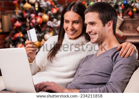 Taking advantages of Christmas sales. Beautiful young couple buying online while using computer together with Christmas Tree in the background