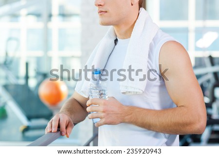 Relaxing after work out. Cropped image of young man carrying towel on shoulders and drinking water while standing in gym