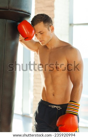 Restoring his energy. Tired young boxer in sports gloves leaning at the punching bag