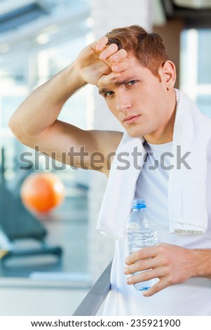 Feeling tired after workout. Tired young man with towel on shoulders looking at camera while holding bottle with water and touching his forehead