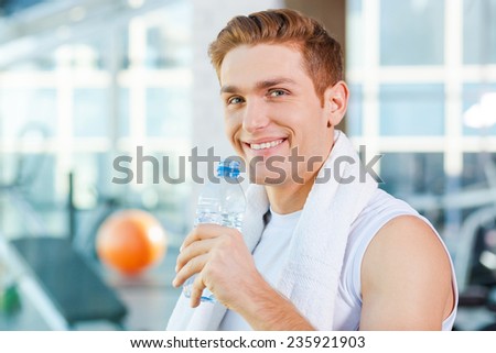Staying hydrated. Handsome young man carrying towel on shoulders and drinking water while standing in gym