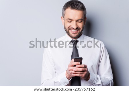 Texting to friend. Confident mature man in shirt and tie holding mobile phone and looking at it with smile while standing against grey background
