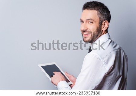 Man with digital tablet. Rear view of happy mature man in shirt and tie working on digital tablet and looking over shoulder while standing against grey background