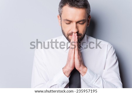 Praying for success. Portrait of concentrated mature man in shirt and tie holding hands clasped near face and keeping eyes closed while standing against grey background