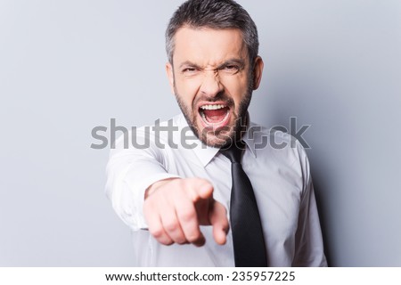 It is your fault! Furious mature man in shirt and tie shouting and pointing you while standing against grey background