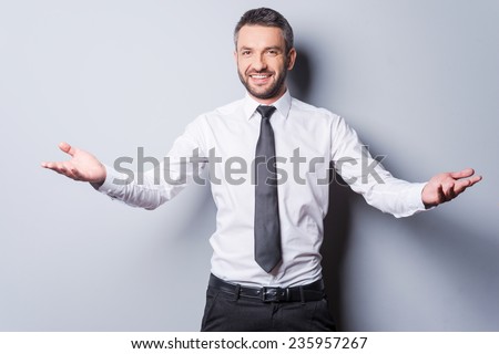 You are welcome! Cheerful mature man in shirt and tie gesturing welcome sign and smiling while standing against grey background
