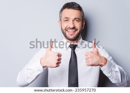 Thumbs up for success! Happy mature man in shirt and tie showing his thumbs up and smiling while standing against grey background