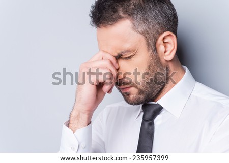 Depressed businessman. Portrait of frustrated mature man in shirt and tie touching his nose and keeping eyes closed while standing against grey backgroundWorried businessman.