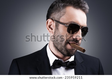 Bossy and self-confident. Portrait of handsome mature man in formalwear and sunglasses smoking cigar and looking away while standing against grey background
