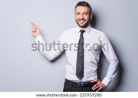 Copy space at his hand. Happy mature man in shirt and tie pointing copy space and smiling while standing against grey background