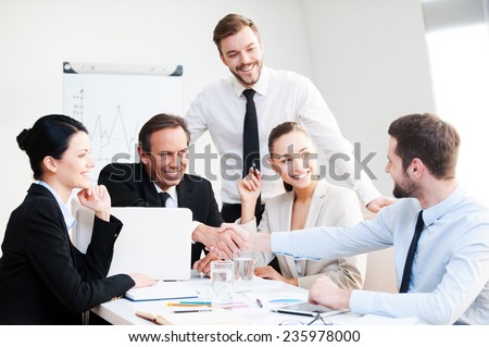 Welcome on board! Group of confident business people in formalwear sitting at the table together and smiling while two men handshaking