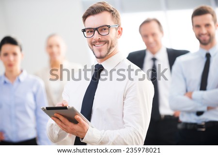 Confident business expert. Confident young businessman holding digital tablet and smiling while his colleagues standing in the background