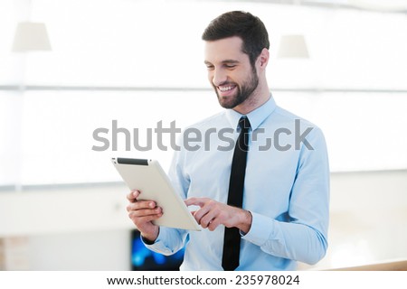 Enjoying work with his new tablet. Handsome young businessman working on digital tablet and smiling while standing indoors