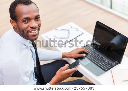 Working with smile. Top view of young African man in formalwear working on laptop and smiling while sitting at his working place