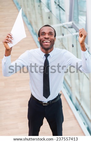 I did it! Top view of happy young African man in shirt and tie holding paper and keeping arms raised