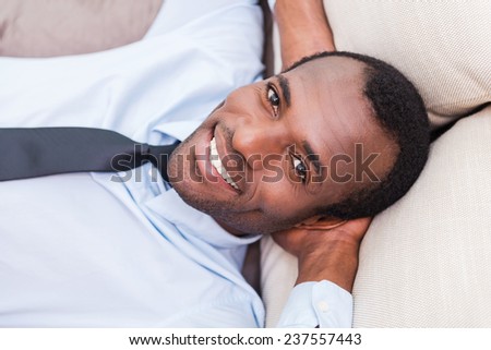 Taking time to for a minute break. Top view of handsome young African man in shirt and tie holding hands behind head and smiling while lying on the couch