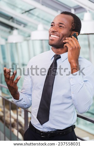 Good business talk. Cheerful young African man in shirt and tie talking on the mobile phone and smiling while standing indoors