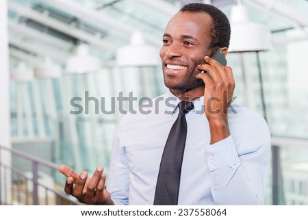 Great news! Happy young African man in shirt and tie talking on the mobile phone and gesturing while standing indoors