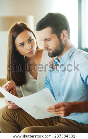 Financial problems. Frustrated young man holding papers and looking at them while woman sitting close to him and pointing document