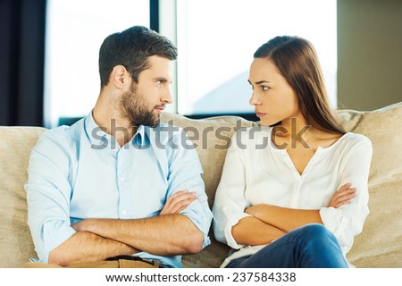 It is your fault! Angry young couple looking at each other and keeping arms crossed while sitting close to each other on the couch