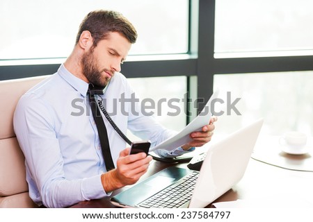 Used to multi-tasking. Concentrated young man in shirt and tie talking on the phone and holding documents while sitting at his working place