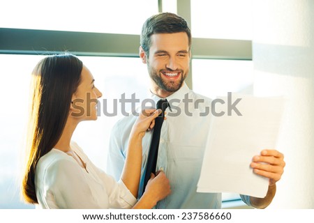 Getting ready to a special day. Handsome young man in shirt and tie holding paper and smiling while woman standing close to him and adjusting his necktie