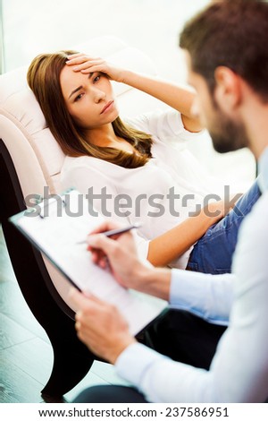 Feeling hopeless and depressed. Depressed young woman sitting at the chair and holding hand on head while young man sitting close to her and writing something in his clipboard