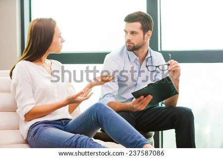Sharing problems with psychiatrist. Worried young woman sitting at the chair and gesturing while male psychiatrist sitting close to her and holding clipboard