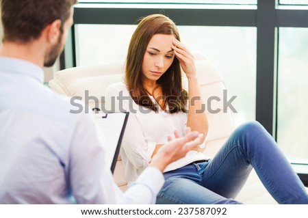 Feeling so hopeless. Depressed young woman sitting at the chair and holding hand on head while male psychiatrist sitting in front of her and holding clipboard