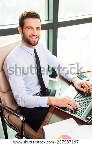 Confident businessman. Top view of handsome young man in shirt and tie looking at camera and smiling while sitting at his working place