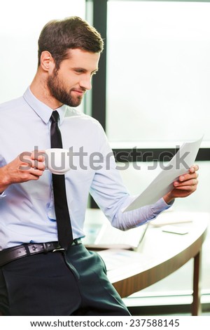 Satisfied with work done. Handsome young man in shirt and tie holding coffee cup and examining document while leaning at the desk