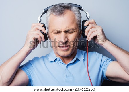 Such music is not for me. Portrait of frustrated senior man in headphones listening to music keeping eyes closed while standing against grey background