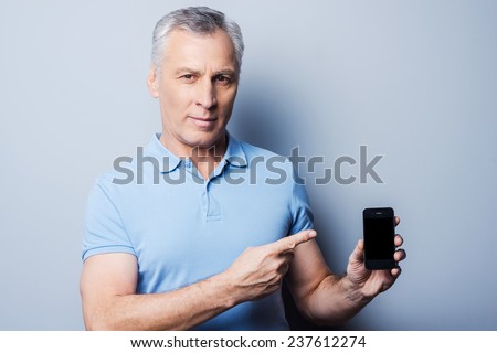Presenting brand new smartphone. Confident senior man in T-shirt showing his new mobile phone and smiling while standing against grey background